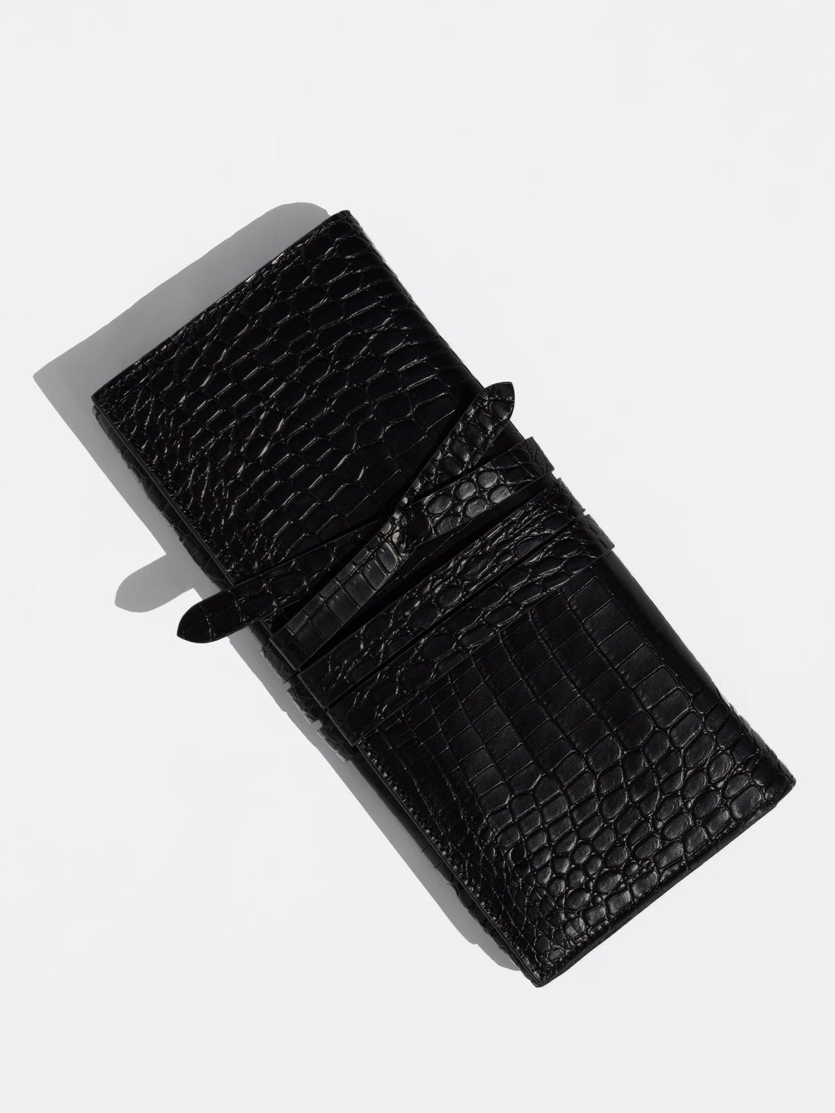 Black crocodile leather clutch bag shown at an angle with its wraparound tie strap, cast shadow on light gray background