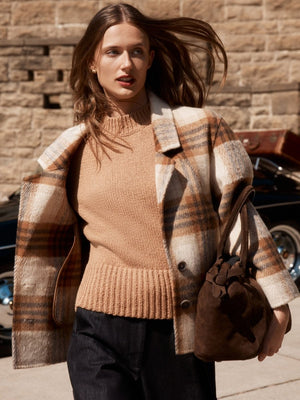 Women's plaid coat in beige and brown tones, styled over a camel knit sweater with dark trousers and a suede handbag.
