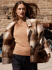 Women's plaid coat in beige and brown tones, styled over a camel knit sweater with dark trousers and a suede handbag.