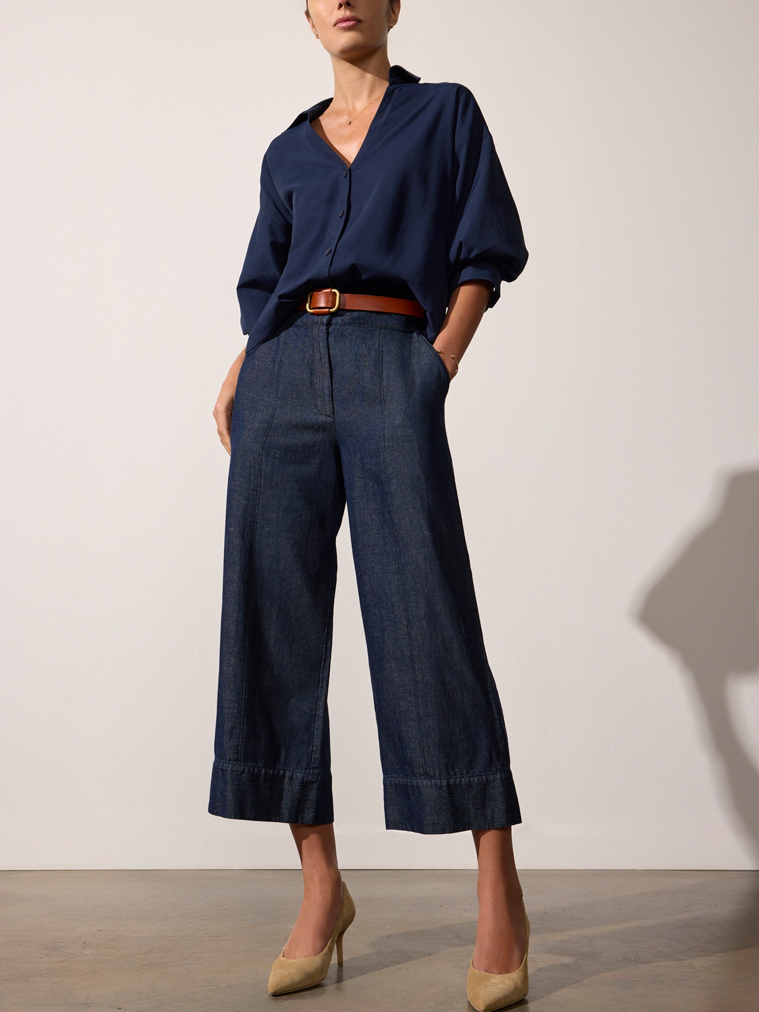 Women's indigo blue pants with wide cropped legs, styled with a navy blouse, tan belt, and beige heels.