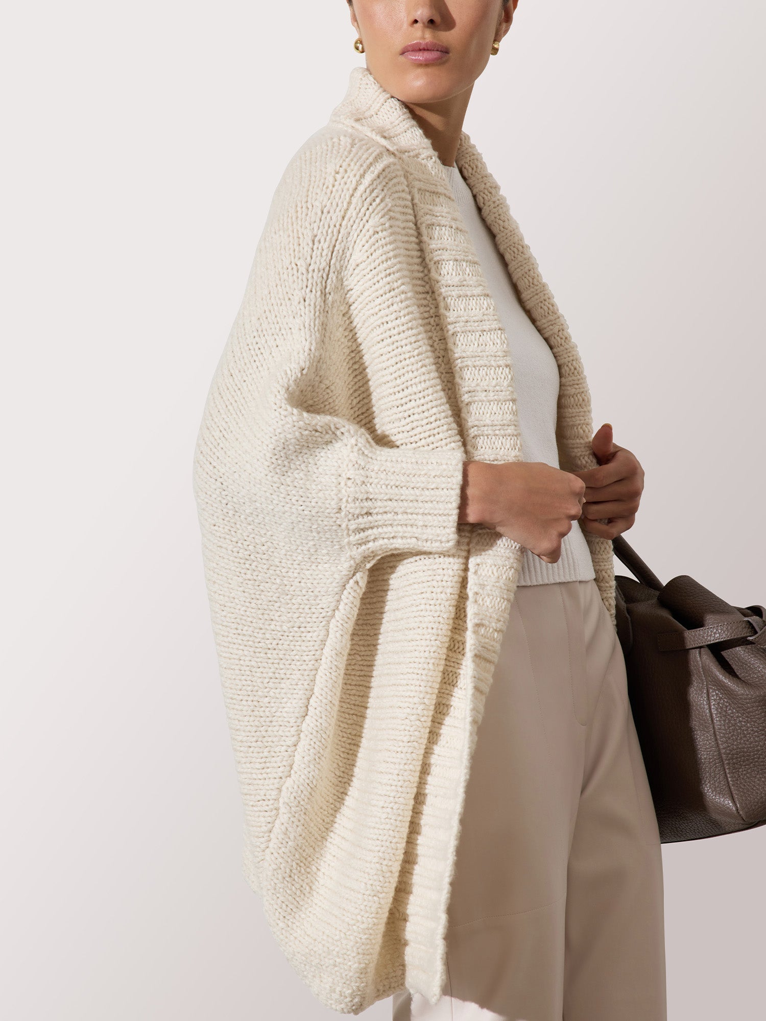 Woman wearing a chunky cream knit cardigan over a white top and beige trousers, carrying a brown leather handbag