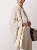 Woman wearing a chunky cream knit cardigan over a white top and beige trousers, carrying a brown leather handbag