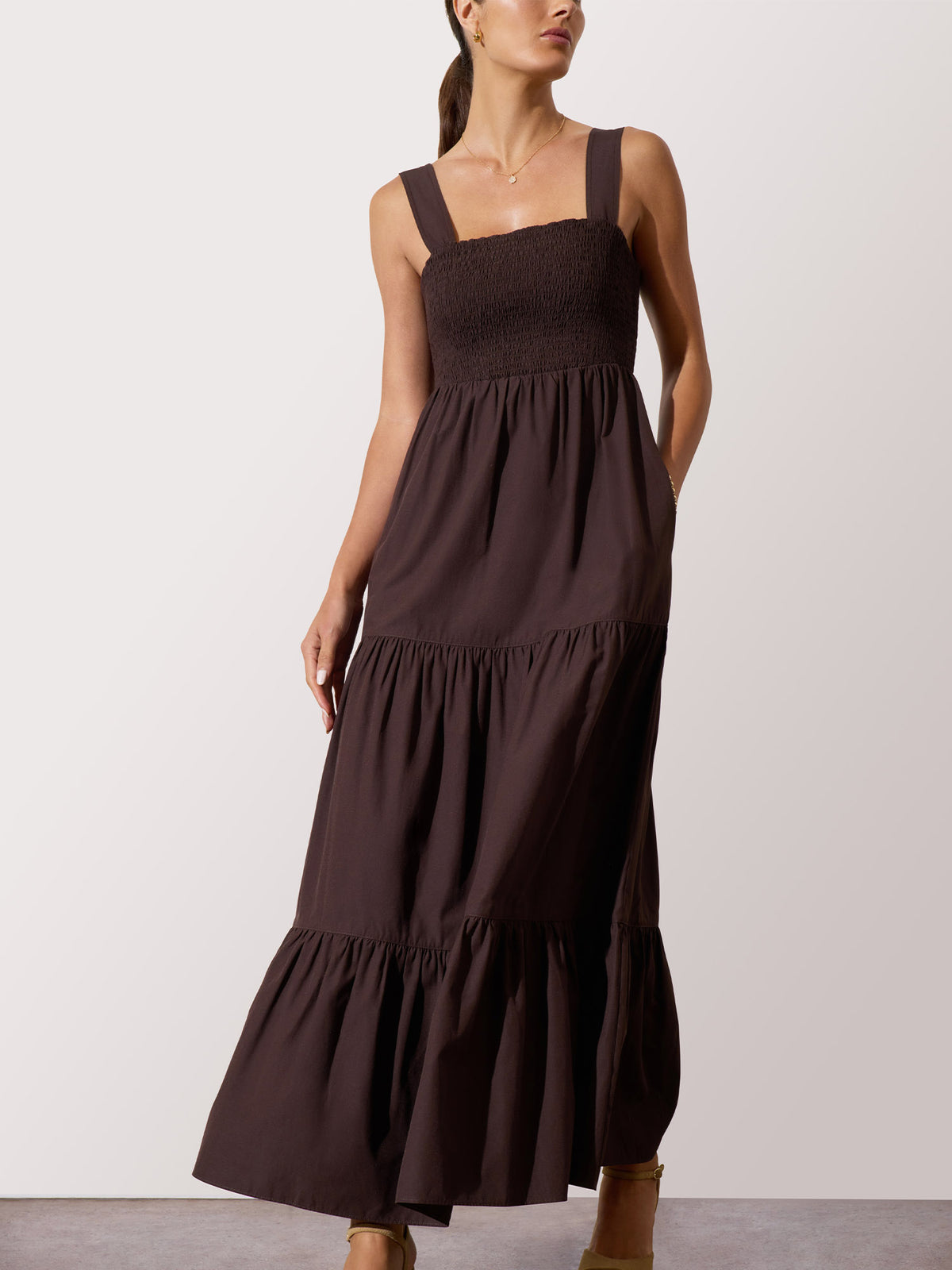 Woman modeling a dark brown tiered maxi dress with a smocked bodice and wide shoulder straps, standing against a light gray studio background.