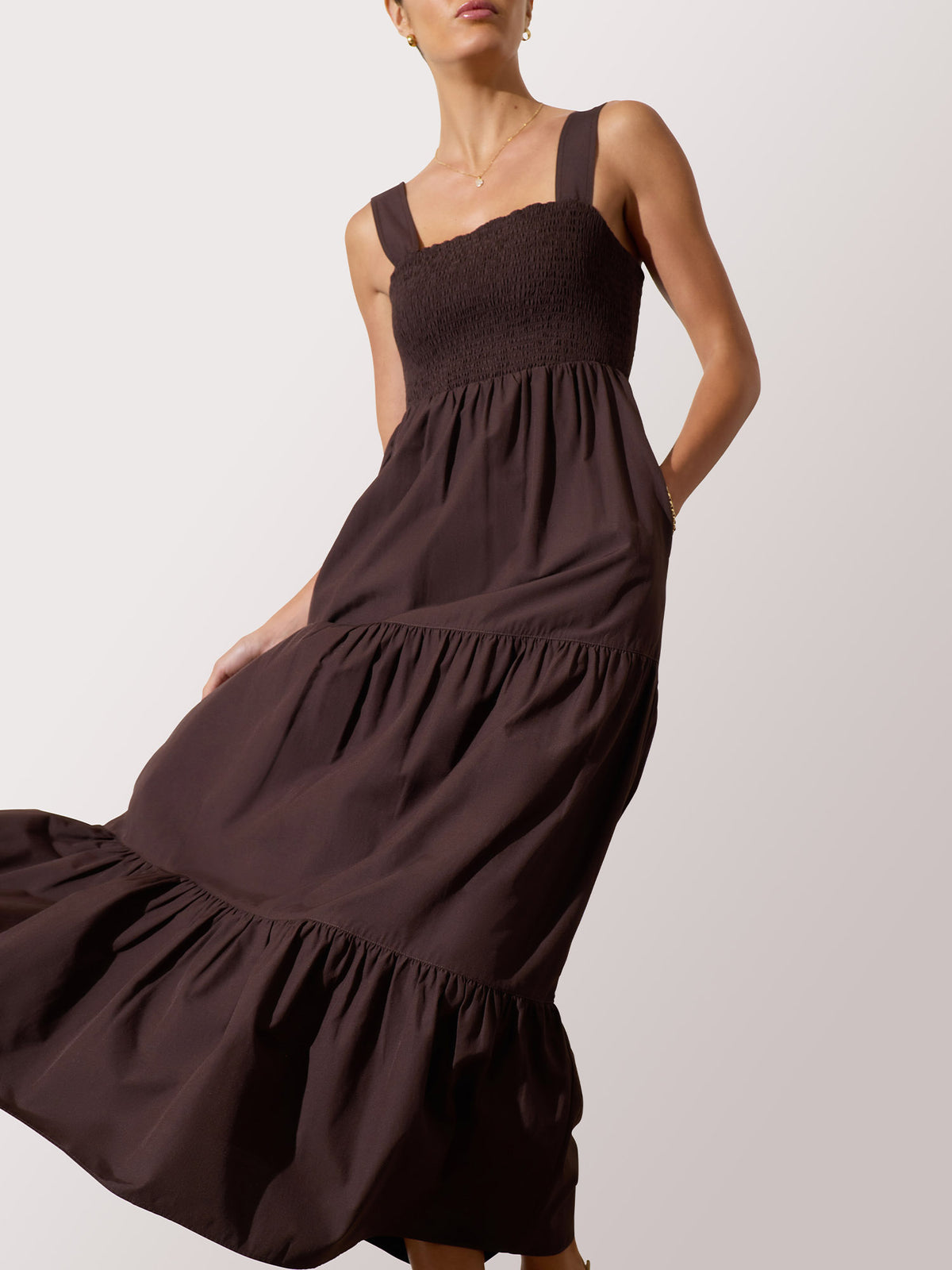 Woman wearing a dark brown tiered maxi dress with a smocked bodice and wide shoulder straps, against a light gray studio background.