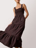 Woman wearing a dark brown tiered maxi dress with a smocked bodice and wide shoulder straps, against a light gray studio background.