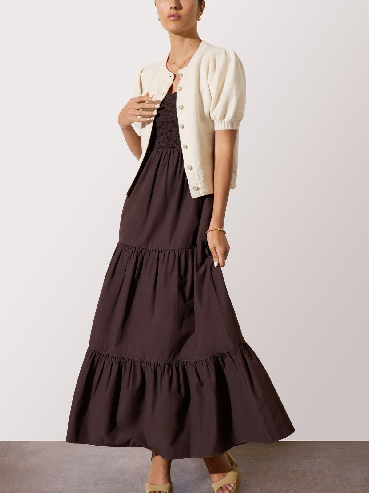 Model wearing a dark brown tiered maxi dress layered with a cream puff-sleeve cardigan, against a light gray studio background.