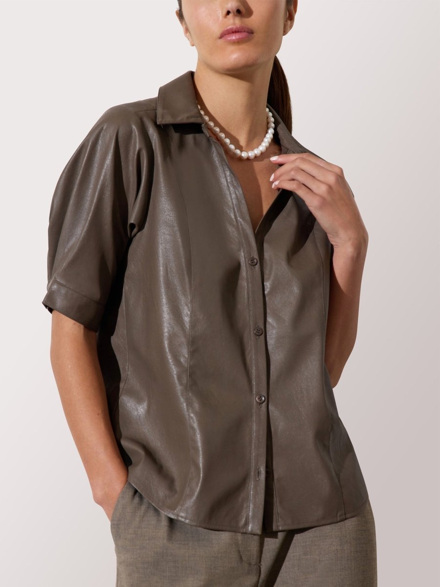 Women's brown leather shirt with short sleeves, collared neckline, and button-down front styled with neutral trousers and pearls.