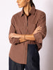 Woman wearing a brown and beige vertically striped button-up shirt with dark blue jeans, against a light gray studio background.