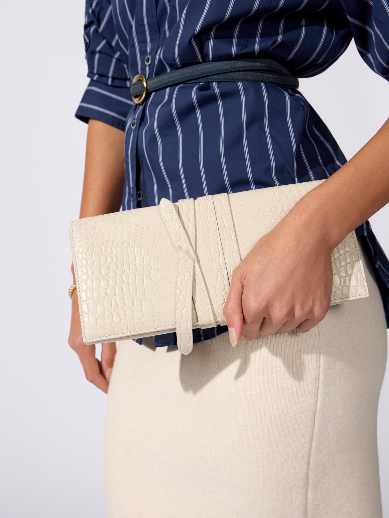 Cream crocodile-embossed clutch bag with a wraparound strap, held against a navy striped shirt and cream skirt.