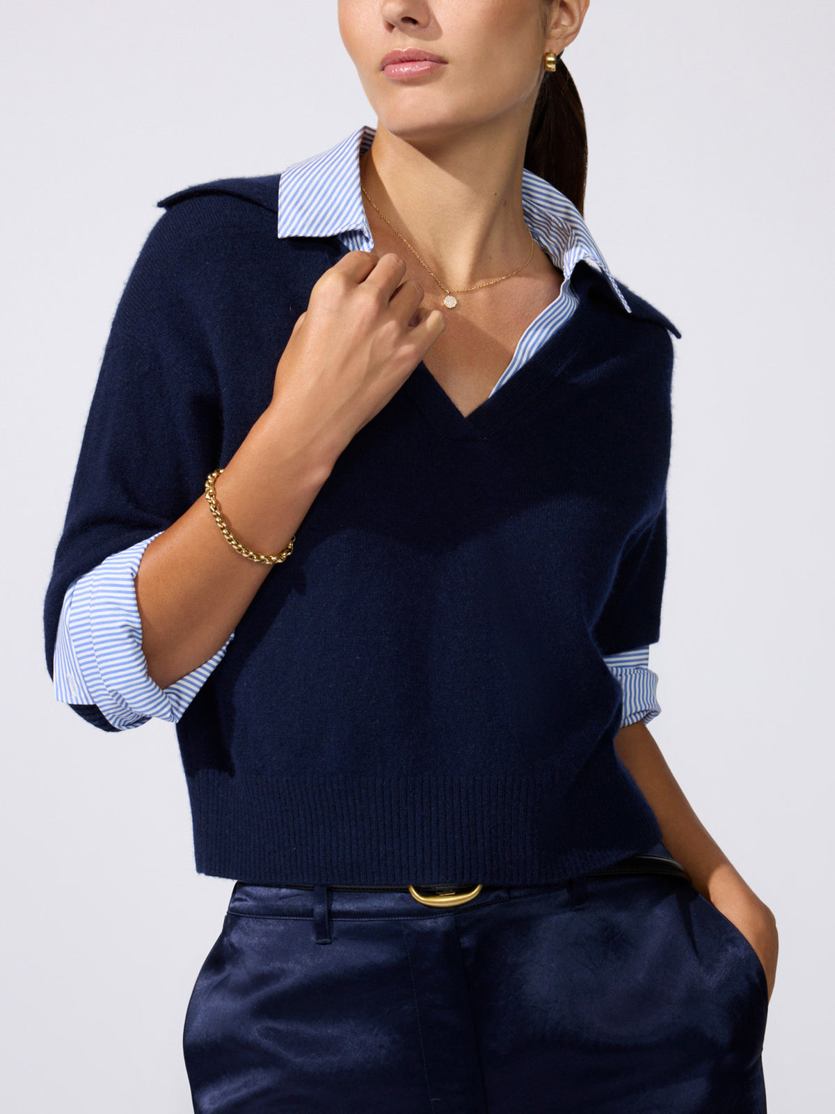 Woman wearing a navy V-neck sweater over a striped shirt with a tan belt, standing by the ocean