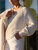 Classy women's sweater in cream chunky cable knit with a V-neckline, balloon sleeves, and ribbed hem worn with white trousers and a cream coat.