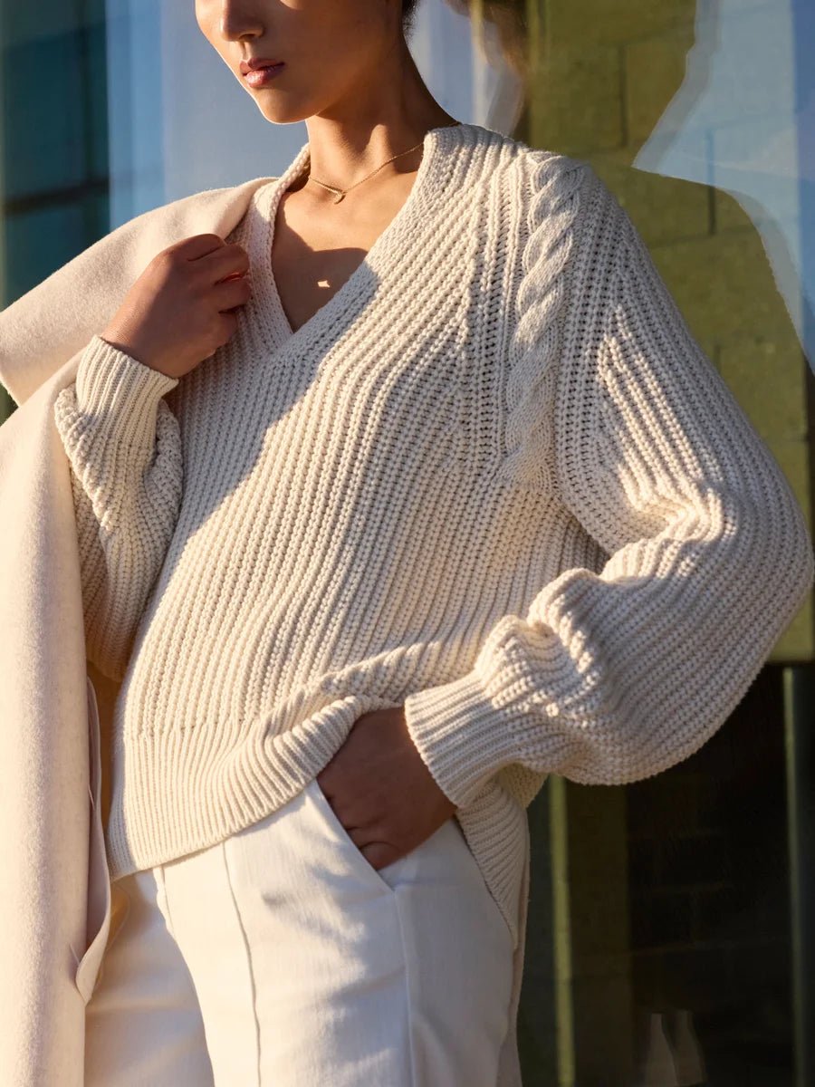 Classy women's sweater in cream chunky cable knit with a V-neckline, balloon sleeves, and ribbed hem worn with white trousers and a cream coat.