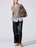 Full-length view of a woman wearing the V-neck pullover in otter melange with flint stripe over a striped shirt, styled with black leather trousers and tan loafers.