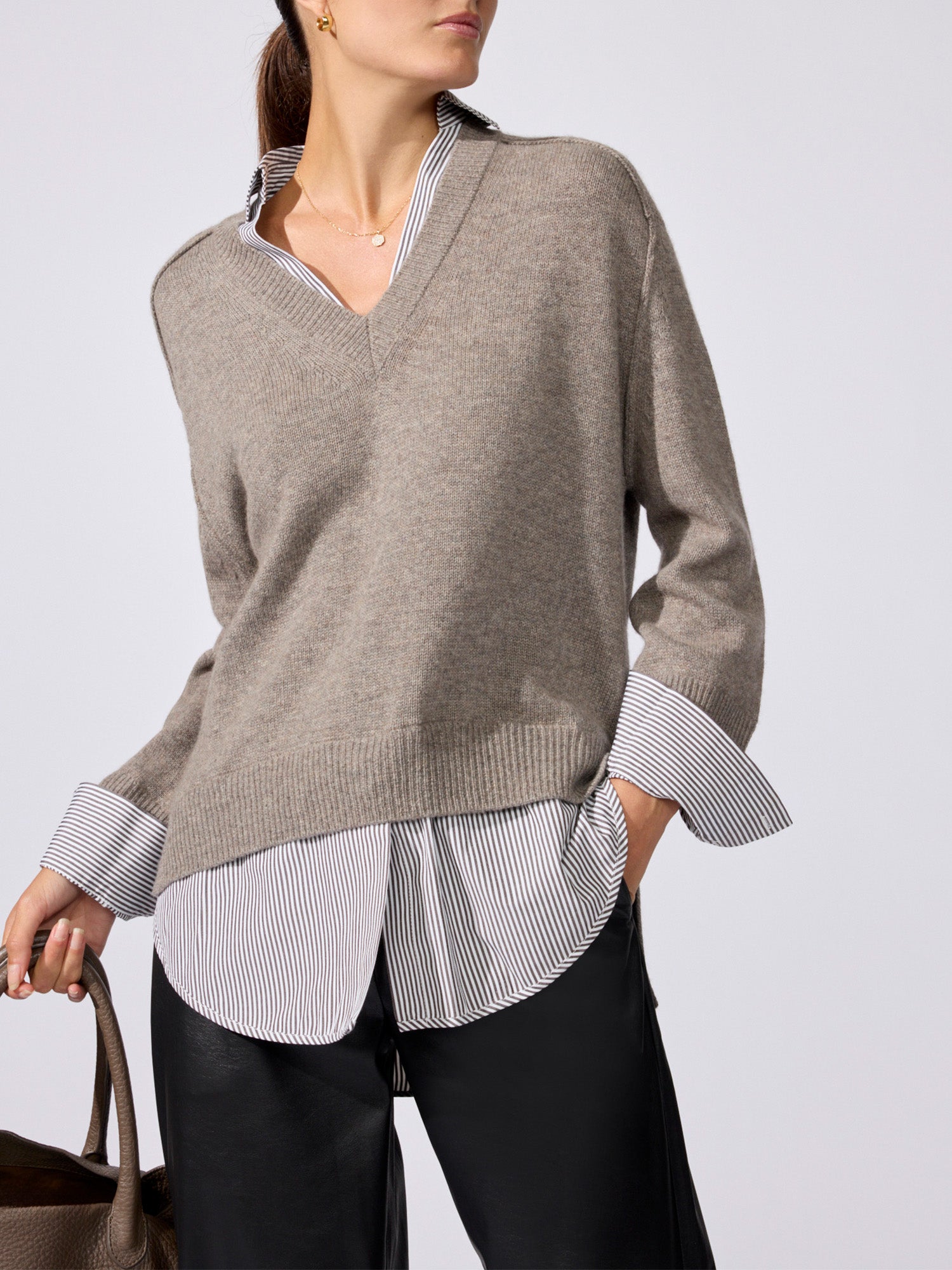 Woman wearing the V-neck pullover in otter melange with flint stripe, layered over a striped button-down with black pants and a leather tote bag.