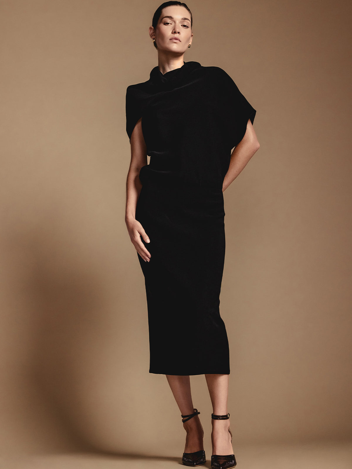 Woman wearing a long black funnel neck dress with draped cap sleeves and black strappy heels against a tan background
