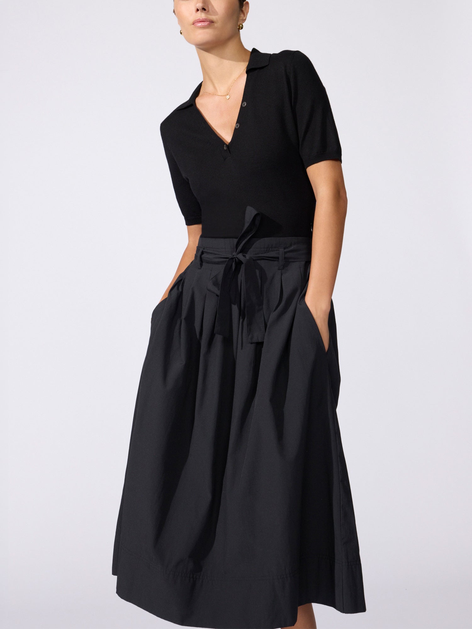 Woman wearing the Isola dress in black onyx, featuring a collared top and a high-waisted pleated skirt with a tie waist