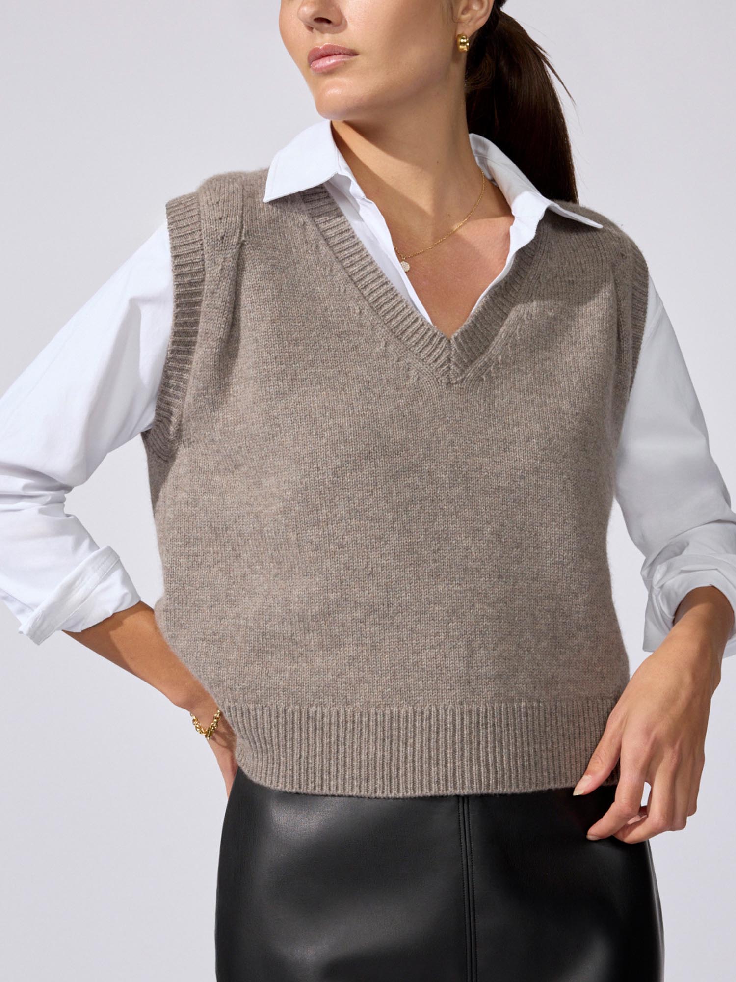 Woman wearing a taupe V-neck knit sweater vest over a white collared shirt, paired with black leather trousers.