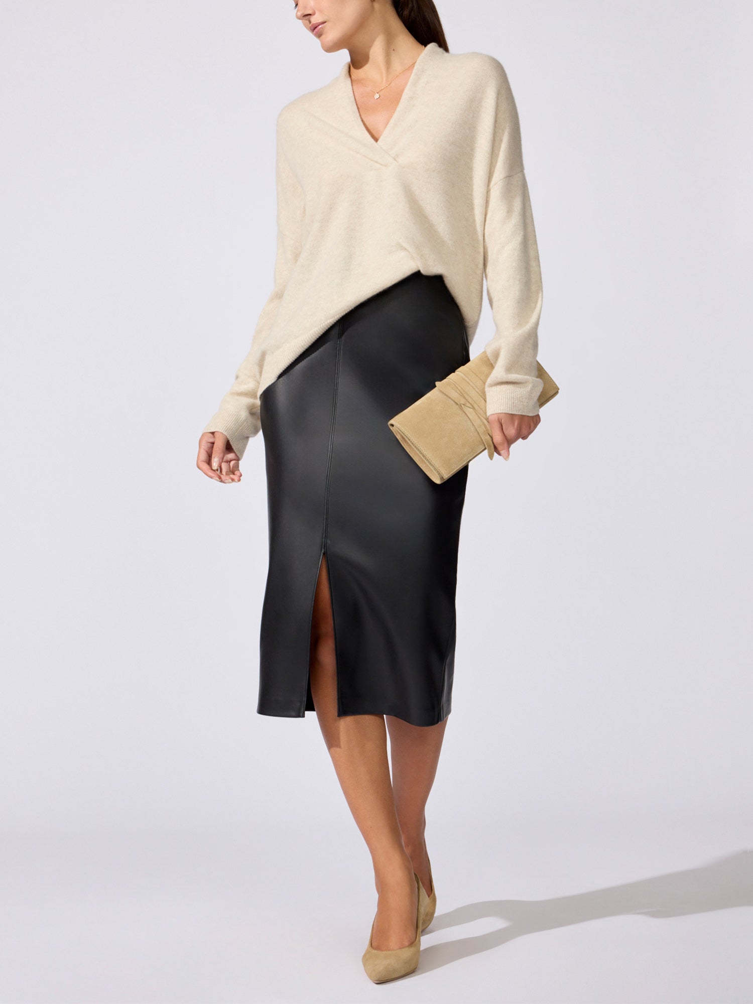 Woman in the Siena wrap-neck cashmere pullover in stone melange with a black leather midi skirt, holding a tan clutch