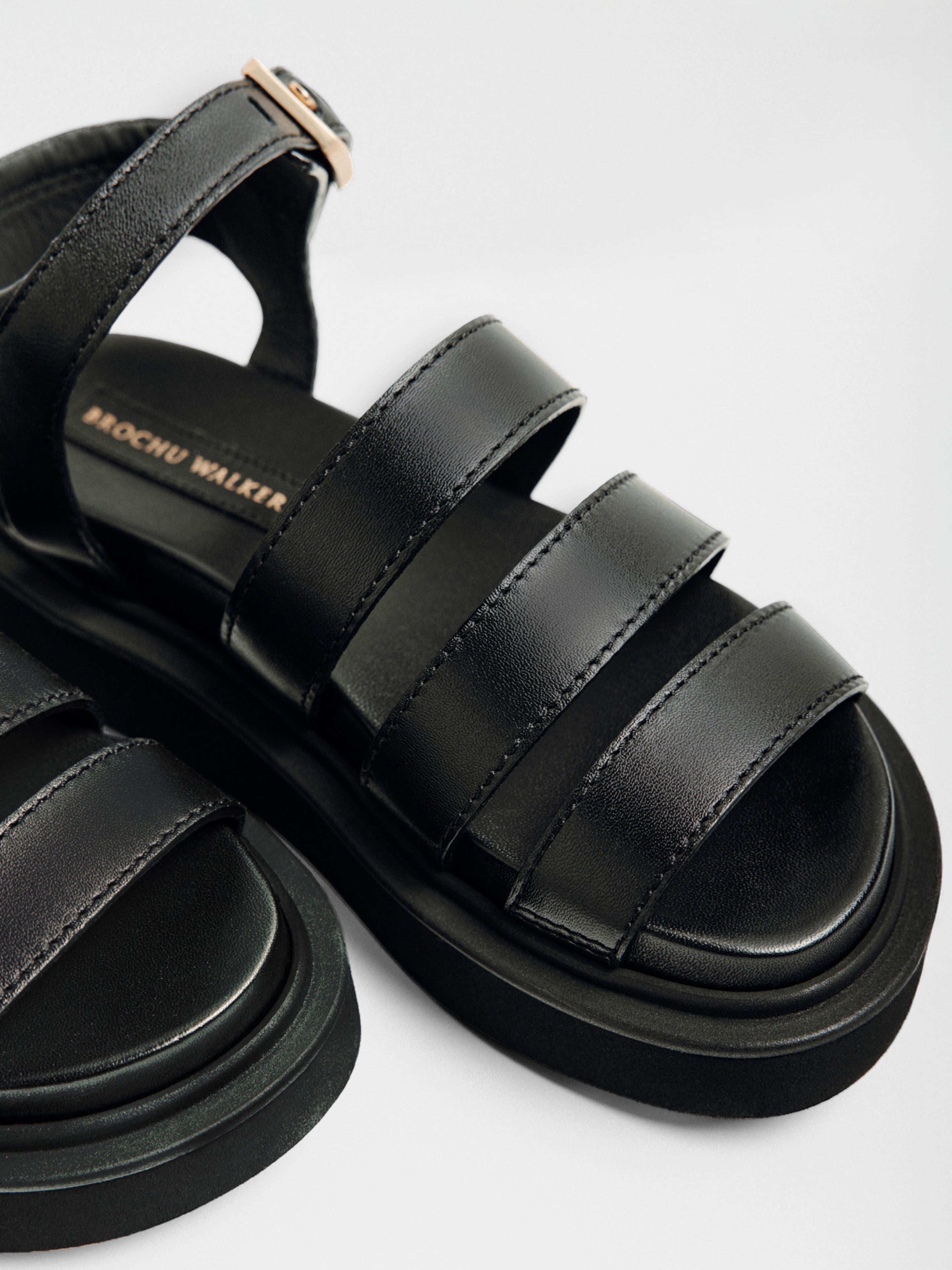 Black sandals with three padded leather straps, a chunky platform sole, and gold ankle buckle shown in a close-up side angle.
