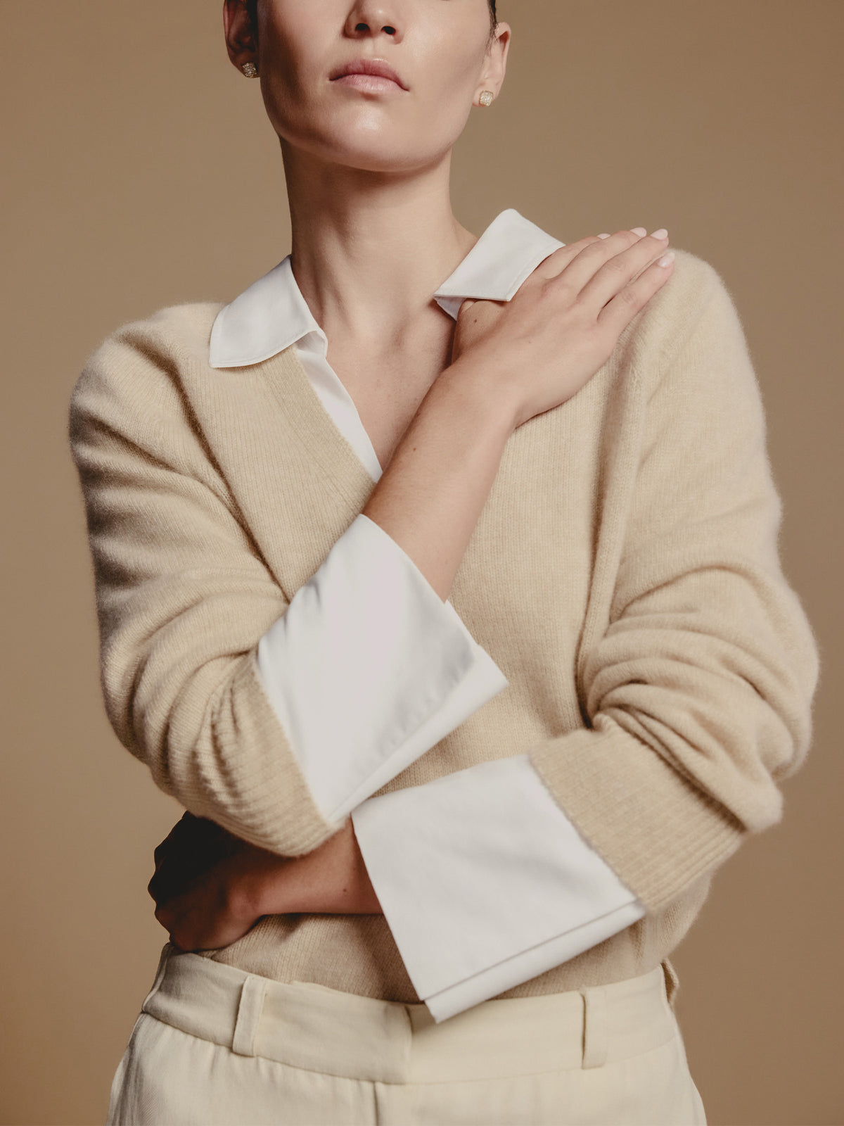 Woman wearing a beige v-neck sweater layered over a white collared shirt with one arm crossed over her chest