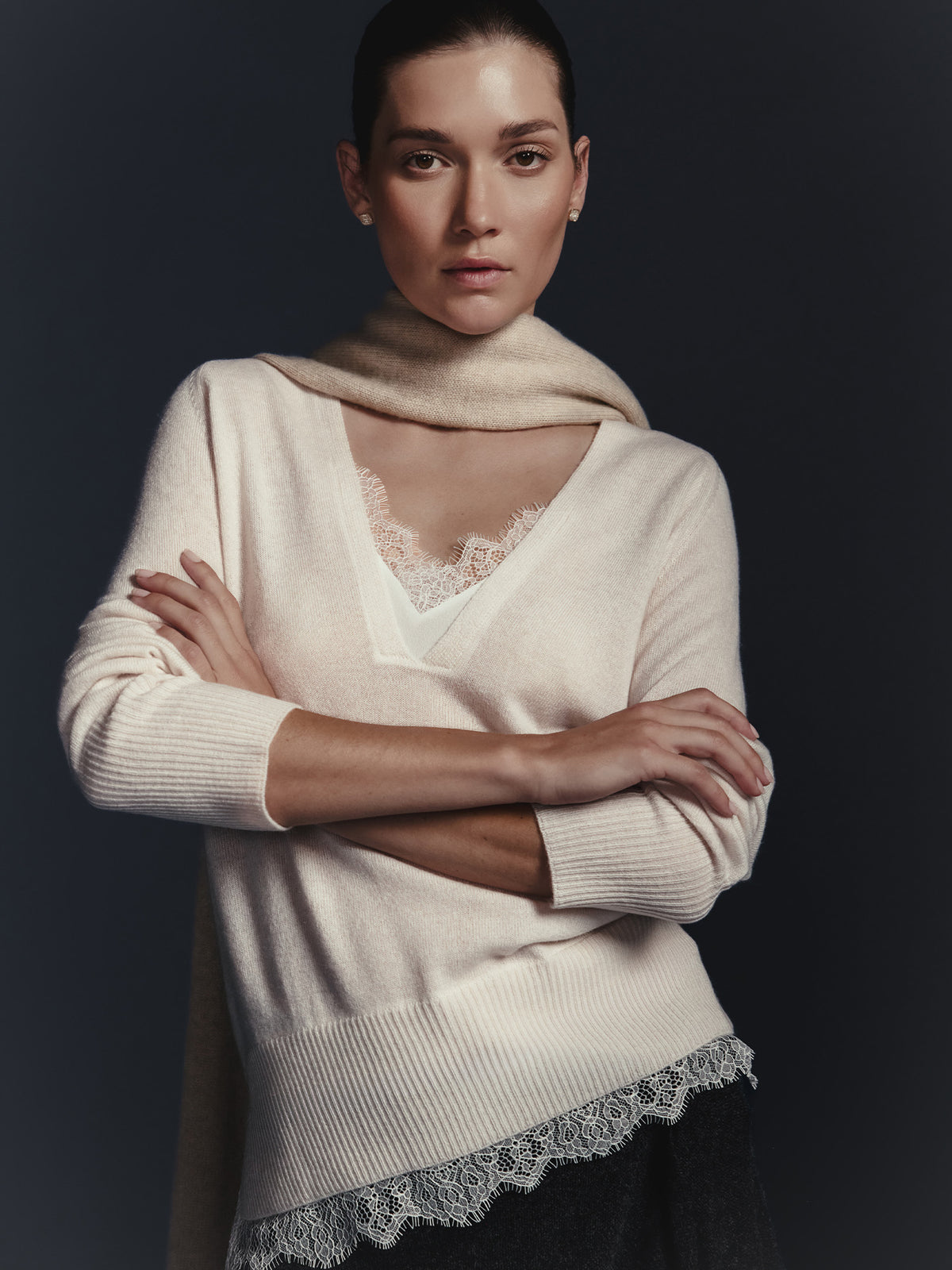 Woman wearing a cream v-neck sweater with a lace trim hem over a lace camisole and a cream scarf against a dark background