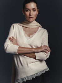 Woman wearing a cream v-neck sweater with a lace trim hem over a lace camisole and a cream scarf against a dark background