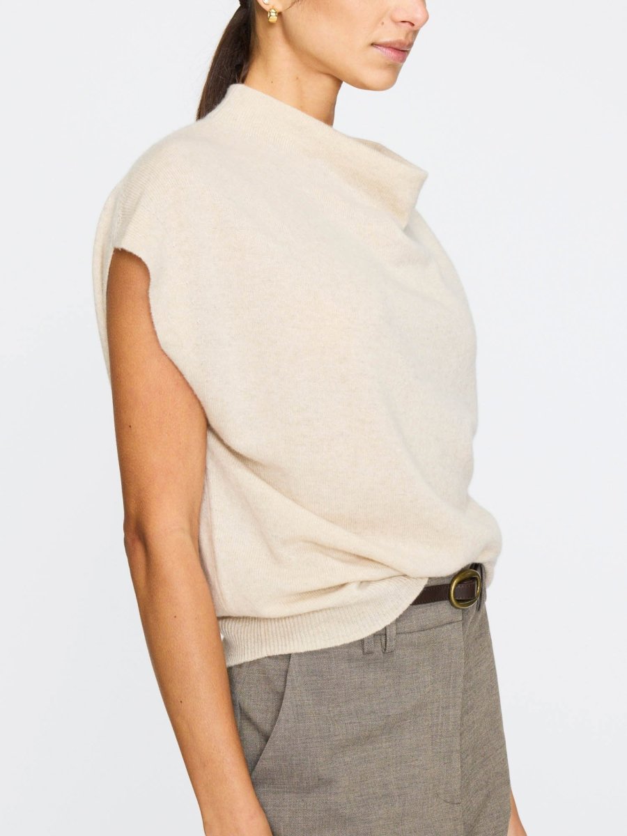 Women's sleeveless cashmere top in a soft beige tone, featuring a draped funnel neck and relaxed fit, styled with tailored trousers.