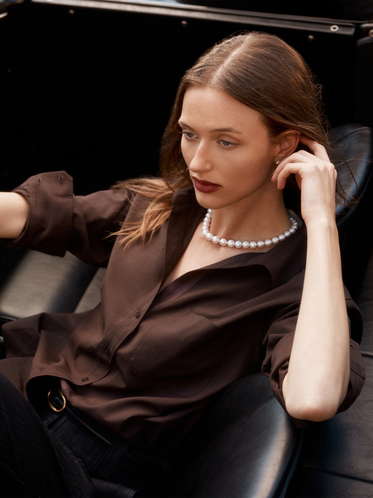 Model in a chocolate brown silk button-up shirt and pearl necklace, seated in a car