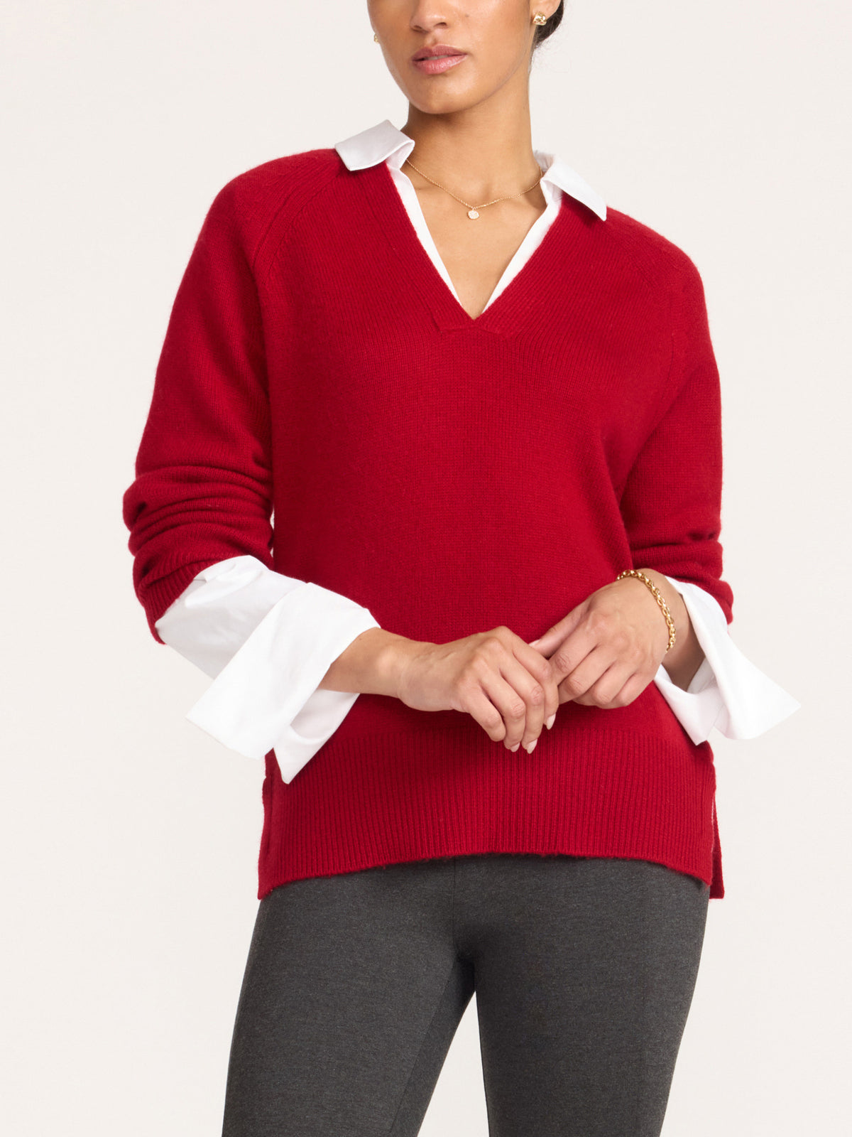 Close-up of a woman in a women's red v-neck sweater with a white shirt collar and flared white cuffs, hands clasped in front of dark gray leggings