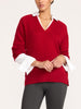 Close-up of a woman in a women's red v-neck sweater with a white shirt collar and flared white cuffs, hands clasped in front of dark gray leggings