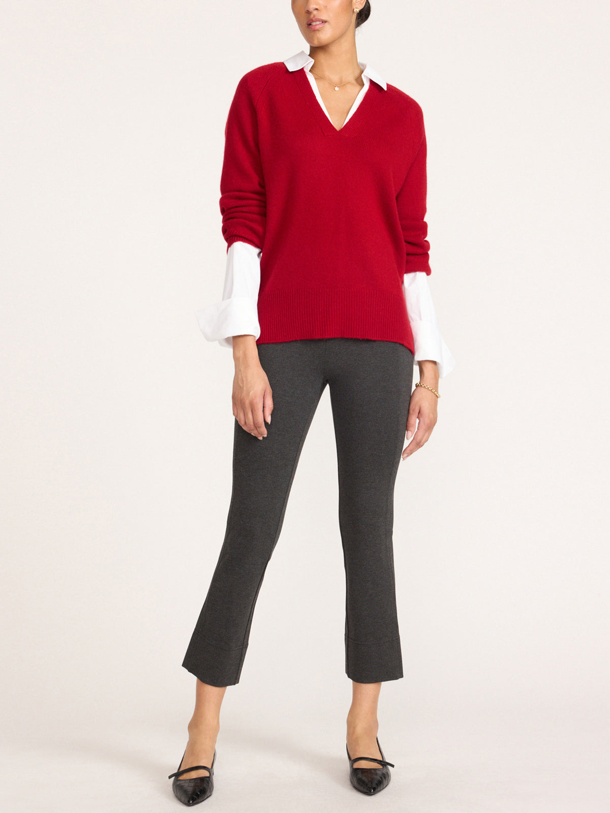 Woman wearing a women's red v-neck sweater layered over a white long-sleeve blouse, with dark gray cropped pants and black pointed flats