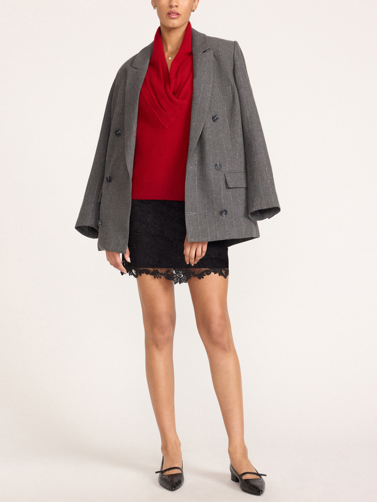 Full-length view of a woman in a women's black lace mini skirt styled with a red cowl neck top, gray double-breasted blazer and black flats