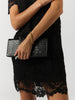Close-up of a women's black lace dress with a scalloped hem, held with a black croc-embossed clutch and a gold chain bracelet