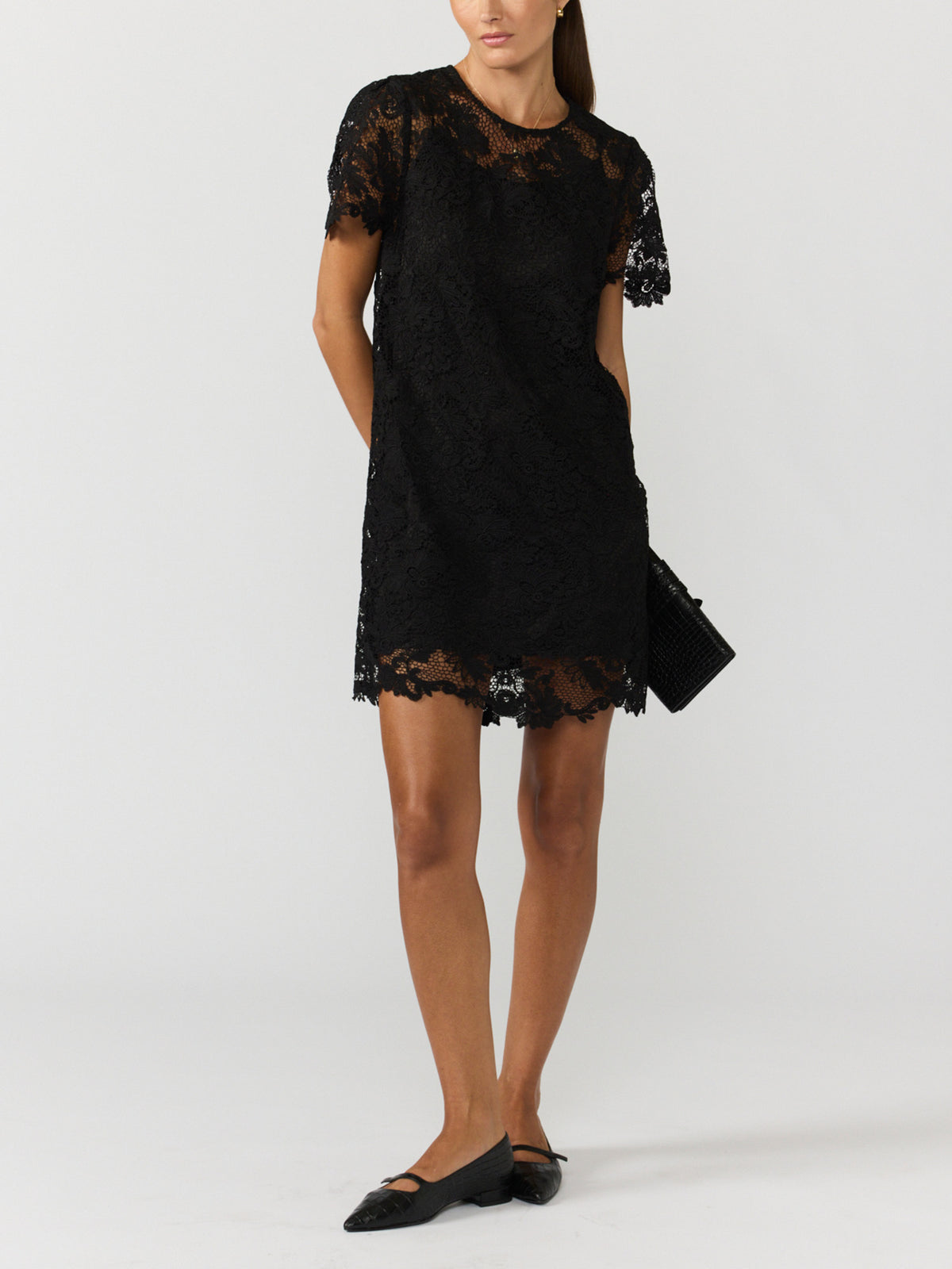 Women's black lace mini dress with short sleeves and a scalloped hem, styled with black pointed flats and a black clutch
