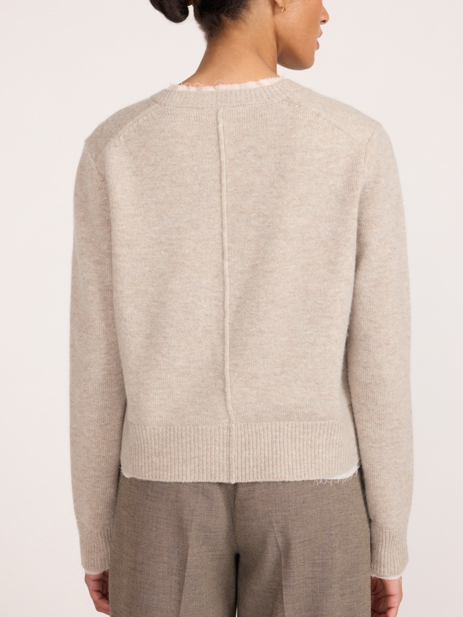 Back view of a woman wearing the Brochu Walker Archie Layered Cardigan in Gravel Stone Melange, a light beige knit sweater with a center seam, paired with taupe trousers.