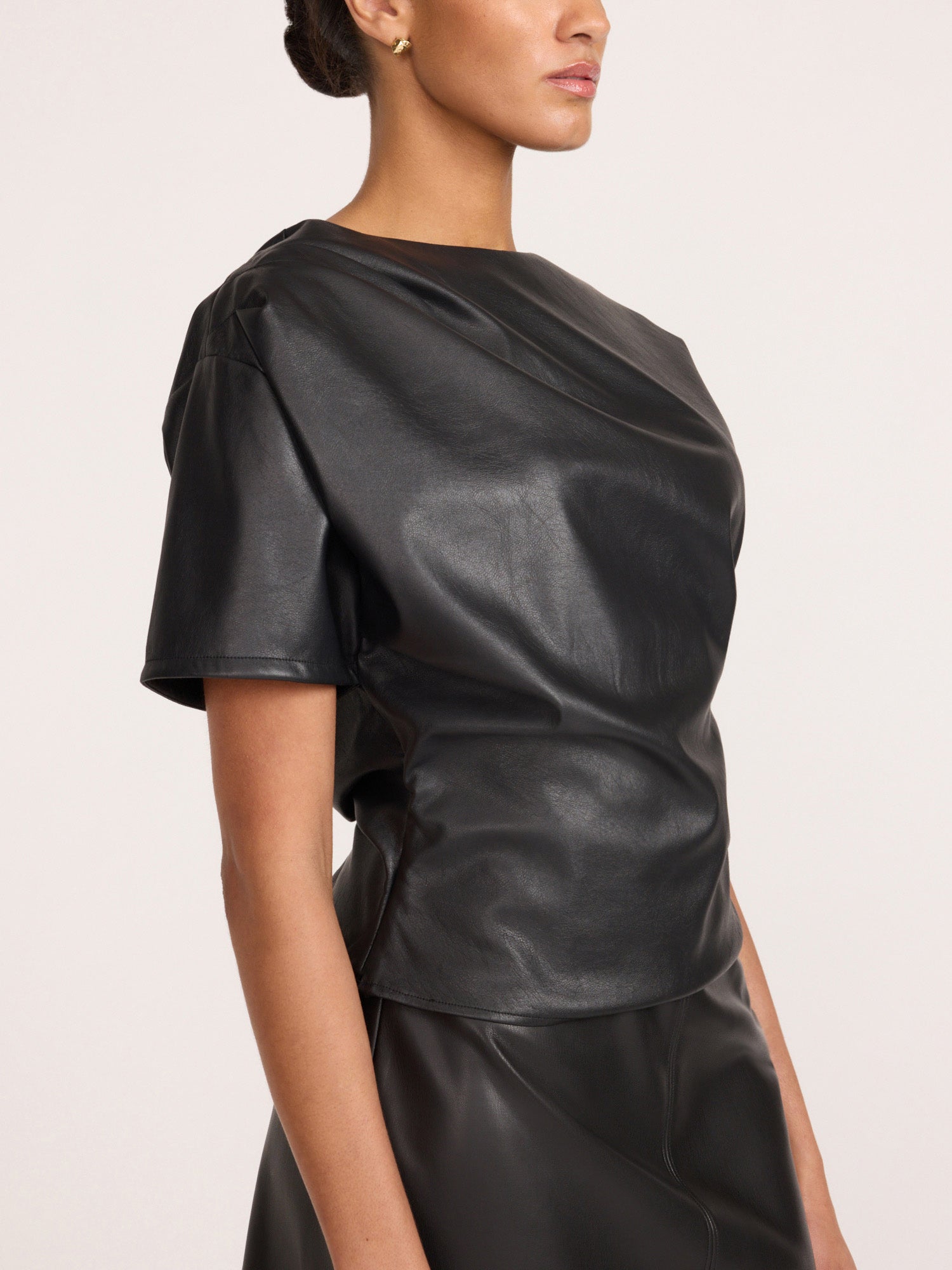 Side view of a woman wearing the Brochu Walker Cleo Top in Black Onyx, a faux-leather top with short puff sleeves, paired with a matching black skirt.