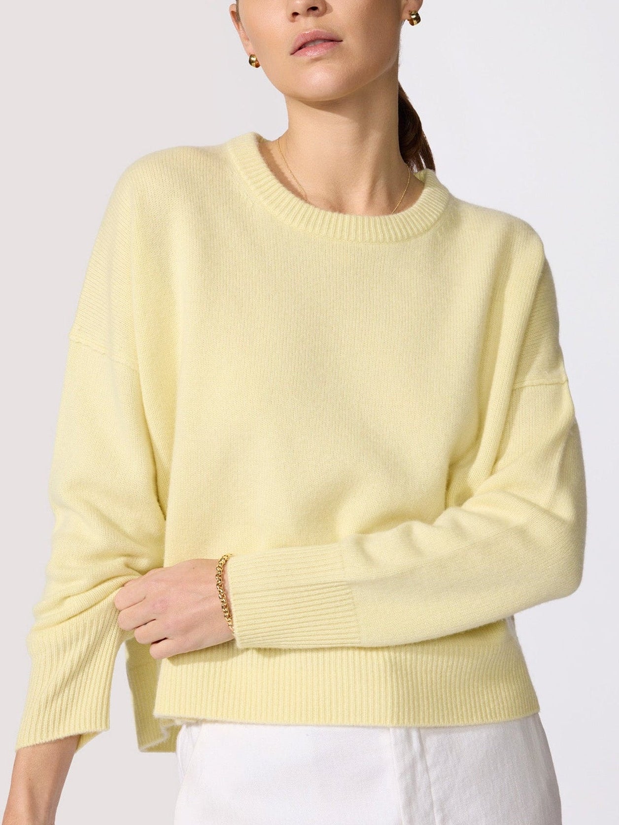 Classy women's sweater in pale yellow cashmere with a crew neck, long sleeves, and ribbed cuffs worn with white trousers shown in close-up.