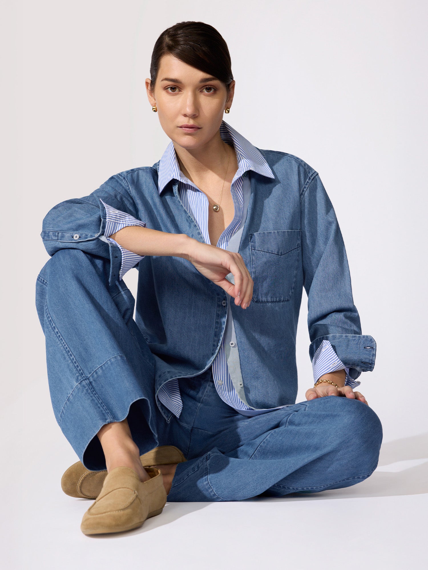 Classy denim outfit in a medium-wash overshirt and wide-leg trousers layered over a striped shirt worn seated on the floor.