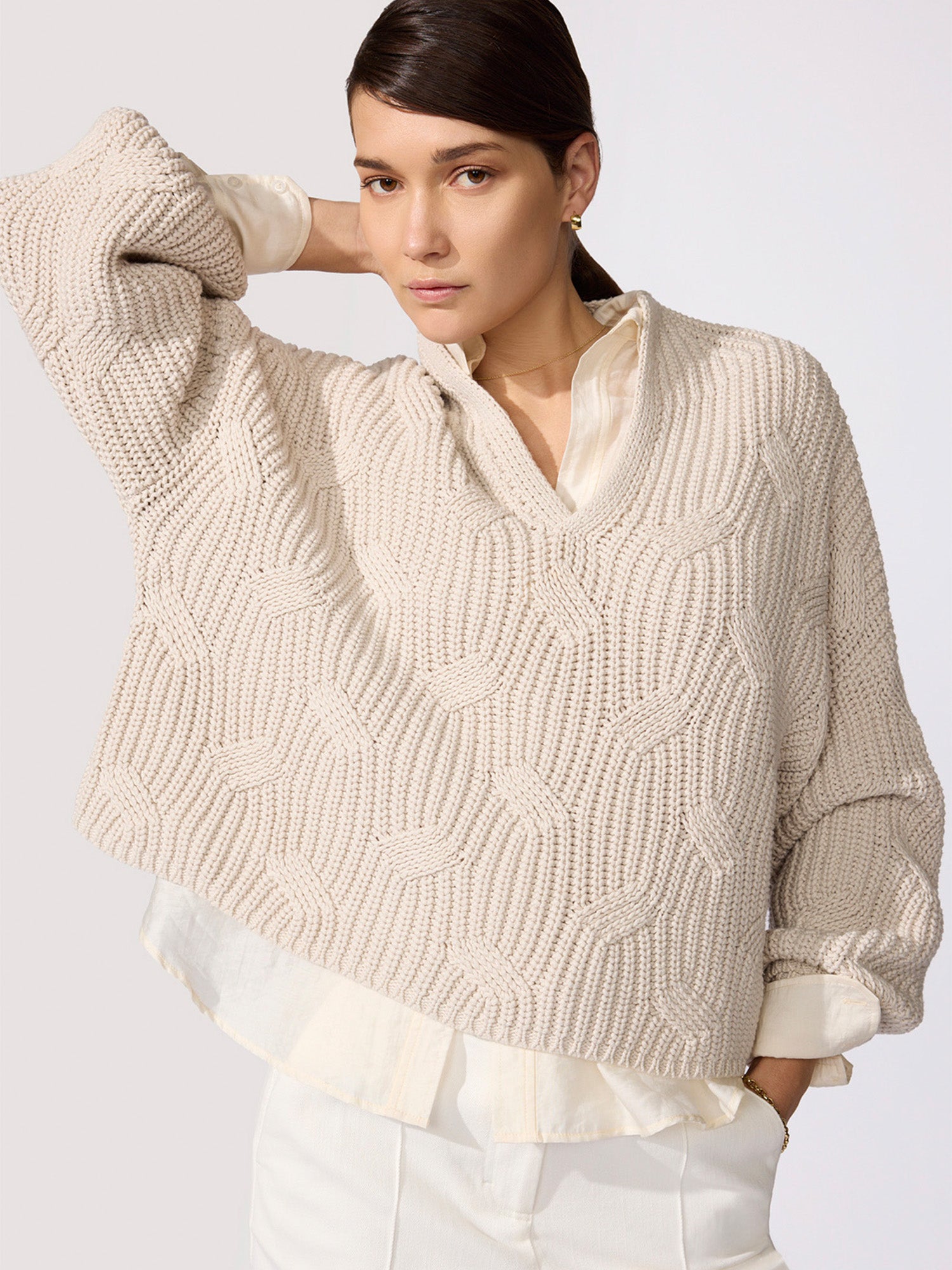 Classy women's sweater in oatmeal chunky cable knit with a V-neckline and wide sleeves layered over a cream shirt with white trousers.