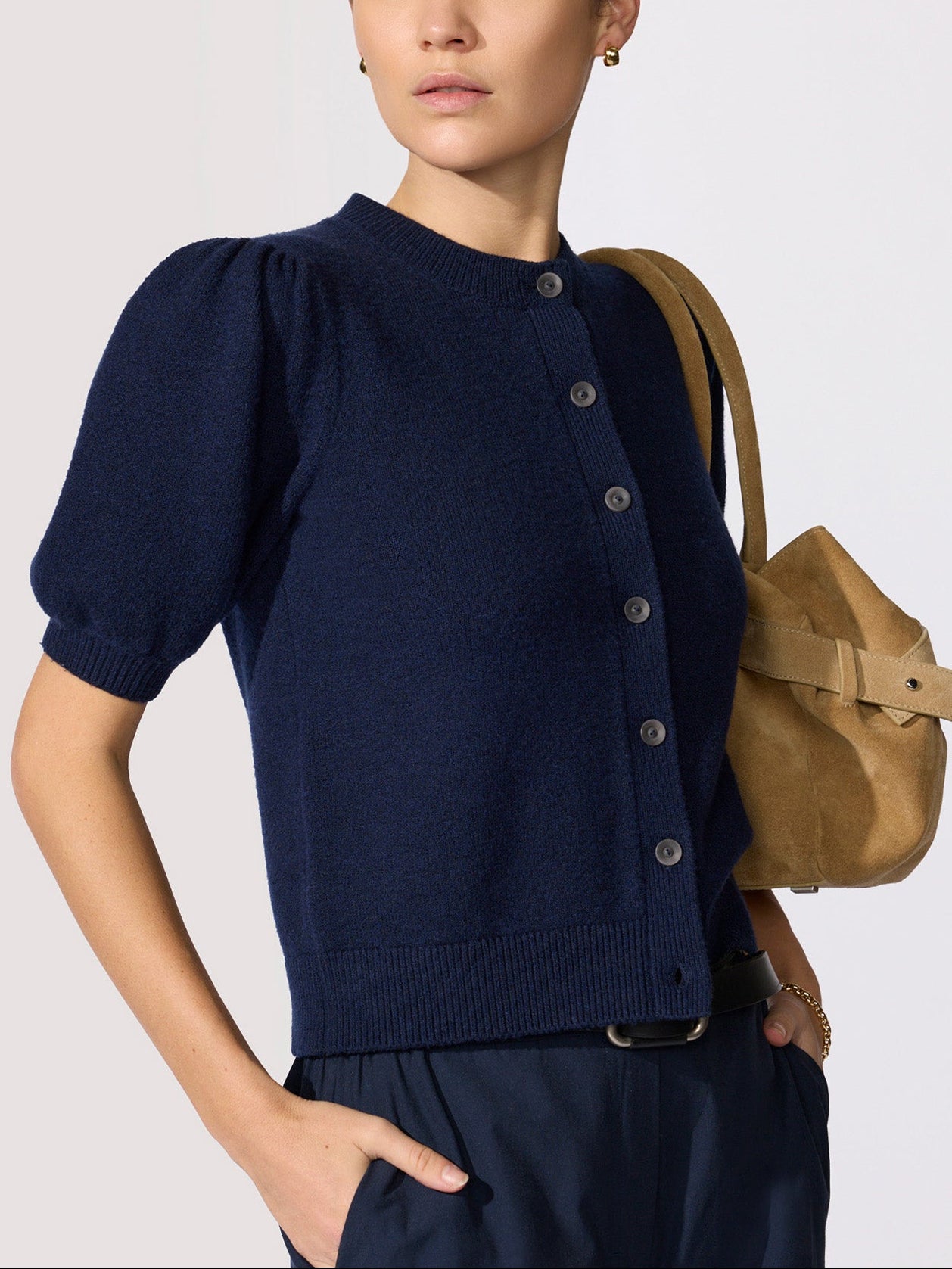 Navy blue outfit close-up with a short-sleeve puff-shoulder button-front cardigan in navy knit worn with a tan suede bag and black belt.