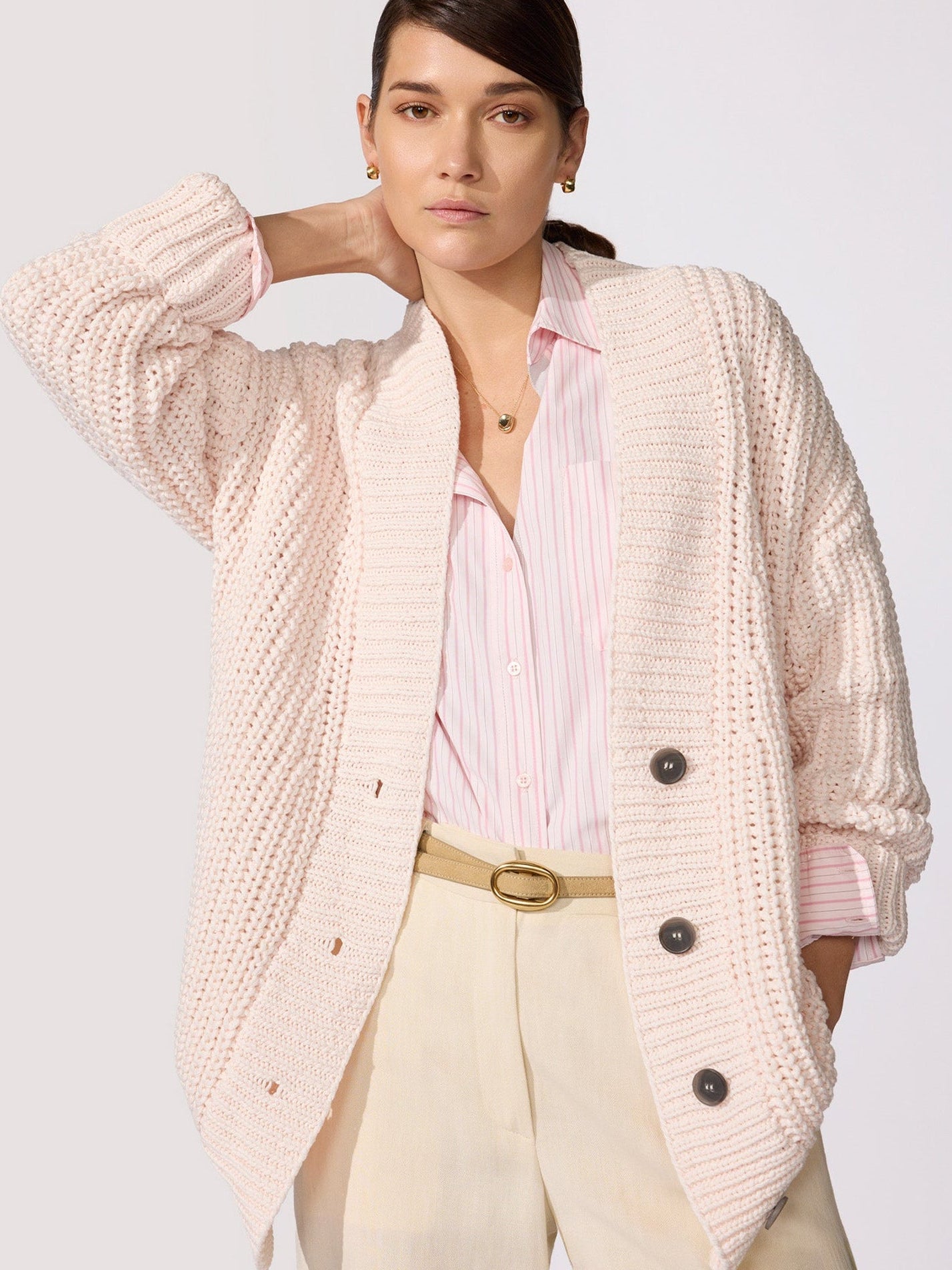 Woman wearing an oversized blush pink chunky knit cardigan layered over a striped button-down shirt with cream trousers.