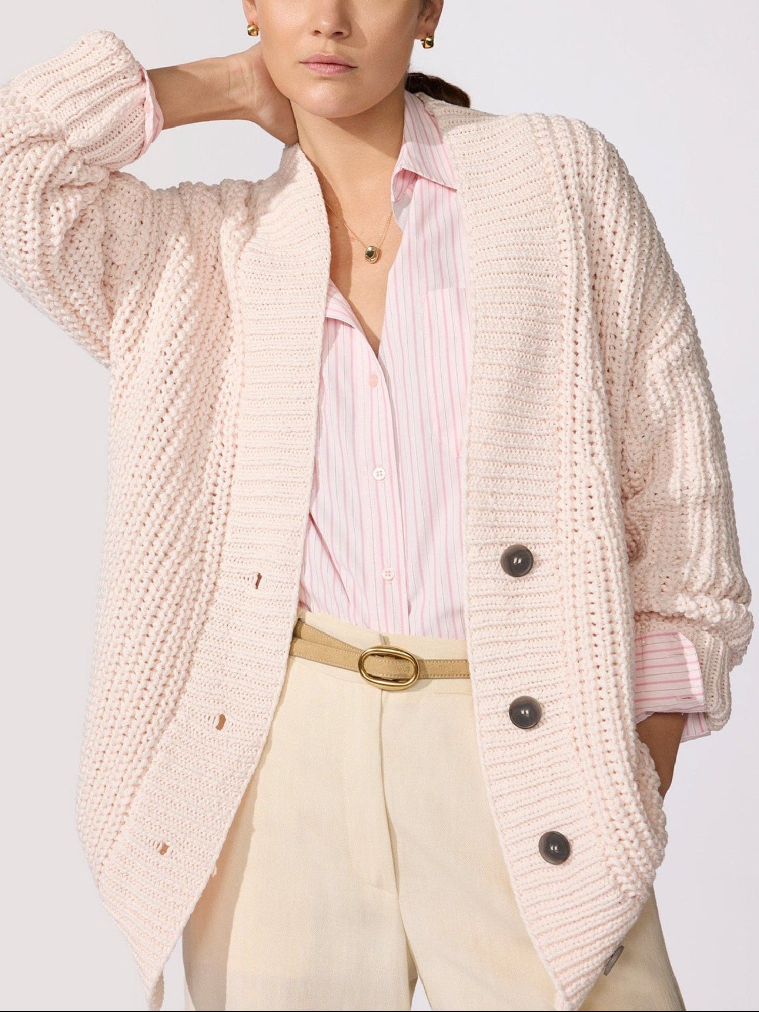 Woman wearing an oversized blush pink chunky knit cardigan layered over a striped button-down shirt with cream trousers.