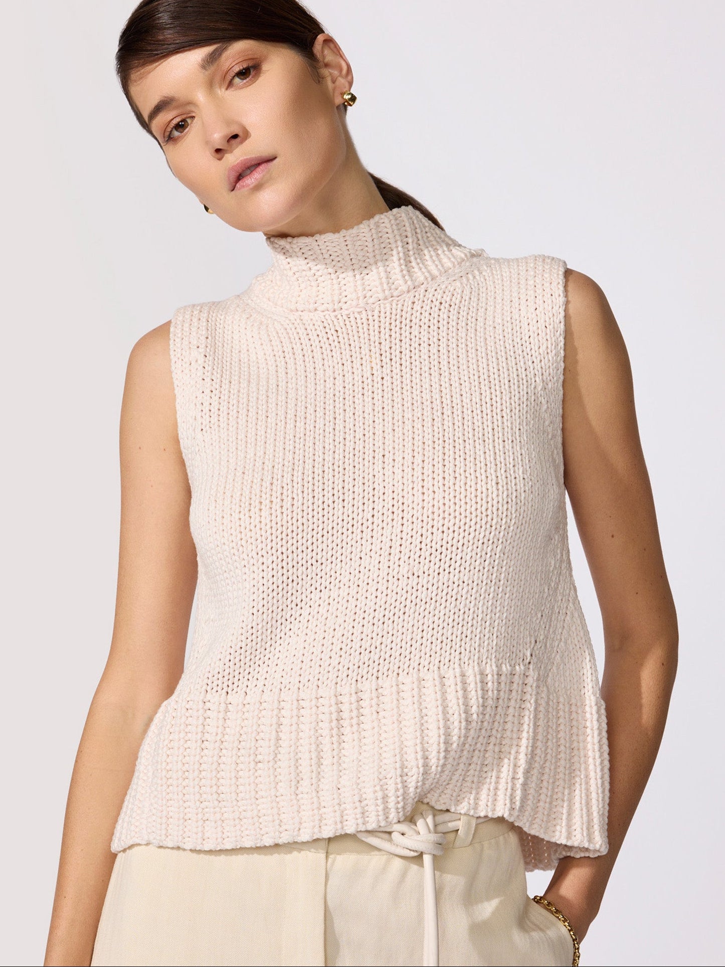 Knitted turtleneck tank top in blush chunky rib knit with a high neck, sleeveless fit, and peplum hem worn with cream drawstring trousers.