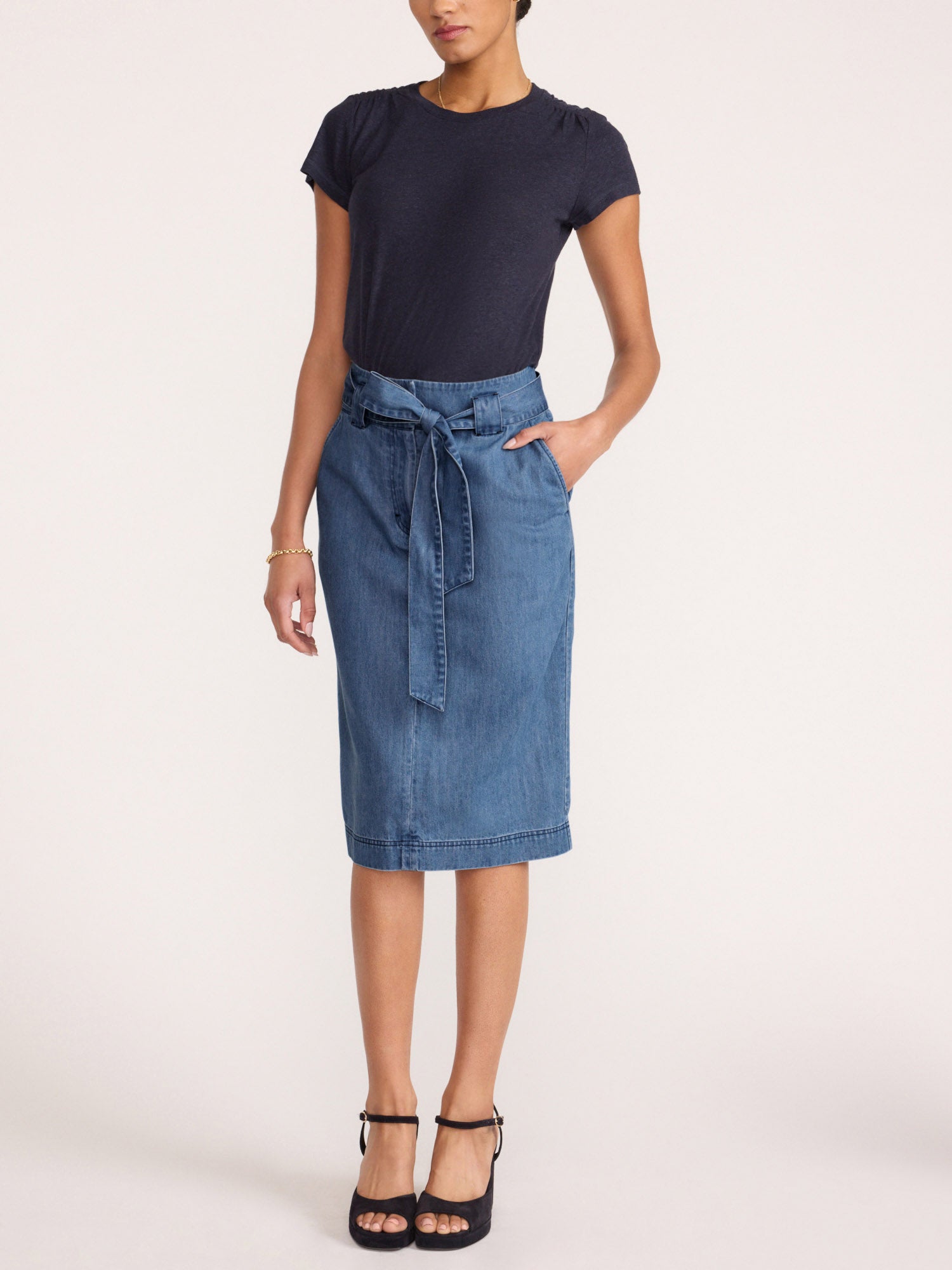 Women's denim skirt in a light-wash midi length with a self-tie waist bow, side pocket, and topstitched hem paired with a navy tee and black heels.