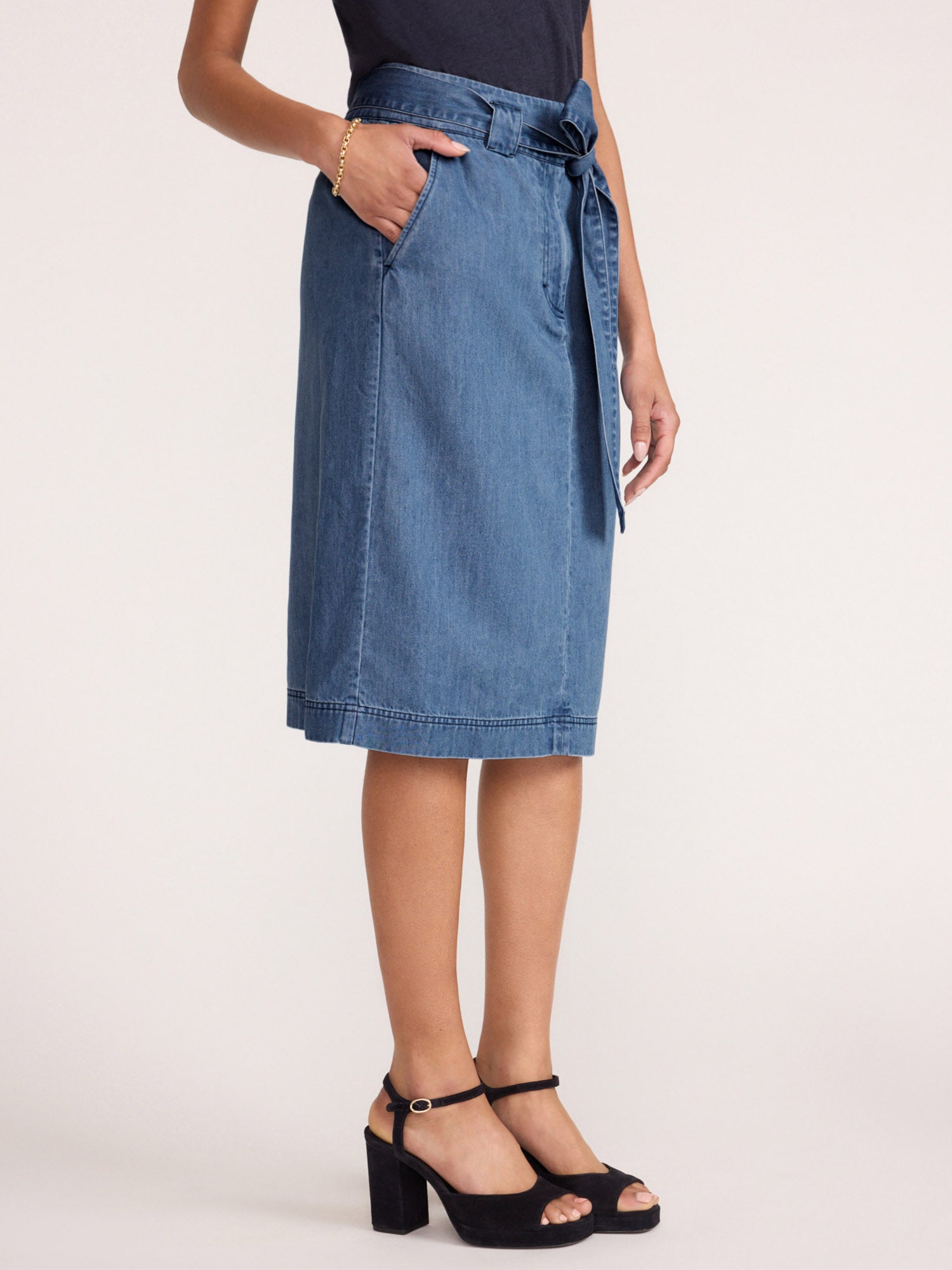 Women's denim skirt in light-wash with a self-tie waist, side pocket, and topstitched hem shown from the side with black platform heels.