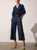 Women's indigo blue pants with wide cropped legs, styled with a navy blouse, tan belt, and beige heels.