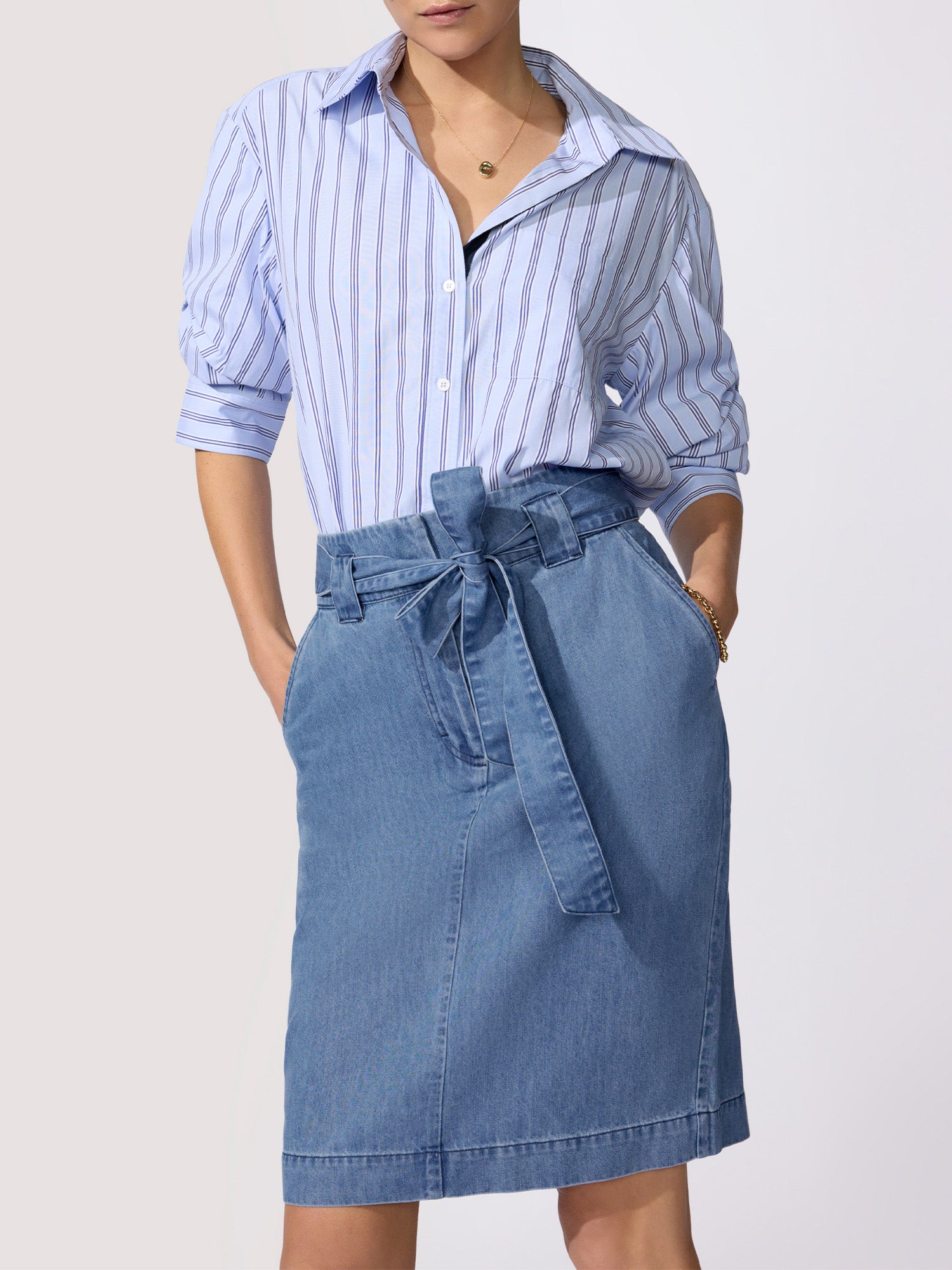 Denim shirt for women in a blue pinstripe worn tucked into a medium-wash denim paper-bag midi skirt with a self-tie bow waist.