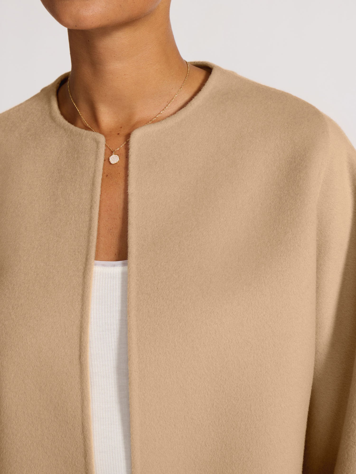 Close-up of a collarless camel wool jacket with a clean open-front neckline, layered over a white ribbed top with a gold pendant necklace.