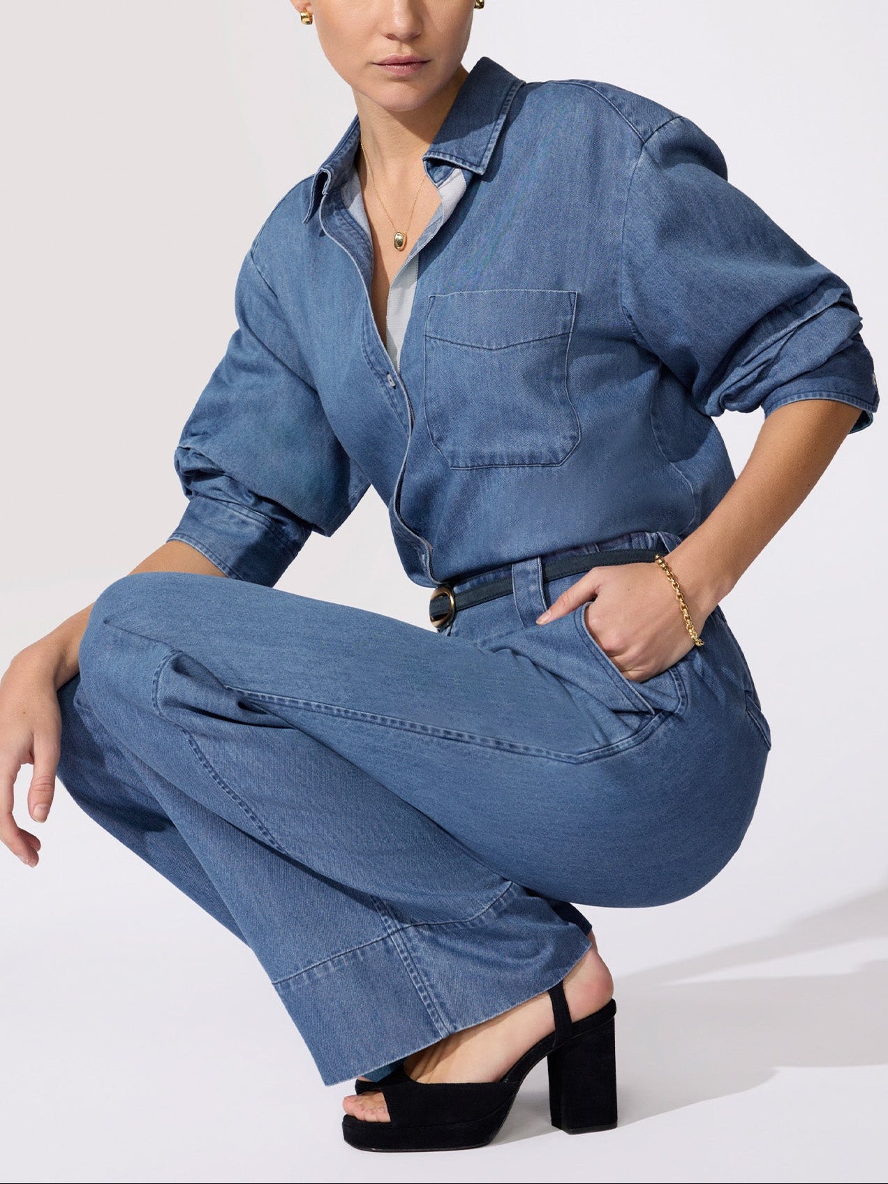 Women's denim outfit with a medium-wash overshirt, chest pocket, and wide-leg trousers with a dark belt worn crouching in black heeled sandals.