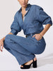Women's denim outfit with a medium-wash overshirt, chest pocket, and wide-leg trousers with a dark belt worn crouching in black heeled sandals.